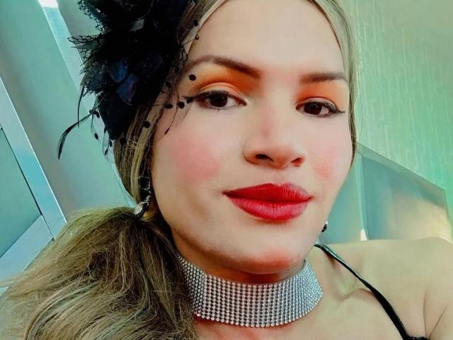Marilyn Marios mit rotem Lippenstift, einem schwarzen Federkopfschmuck und einer glitzernden Halskette, lächelt in die Kamera und zeigt ihr rundes Gesicht und ihren blonden Pferdeschwanz. Ihre Brüste sind unter dem schwarzen Träger sichtbar..