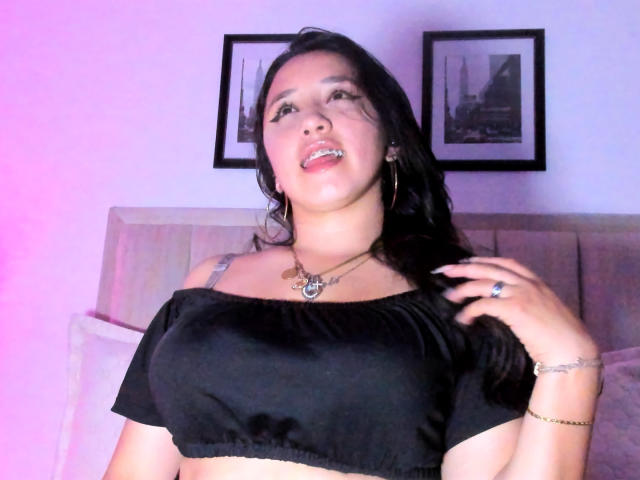 JessyBarnnett, con cabello negro, lengua afuera, vistiendo una blusa negra con un hombro descubierto, mostrando sus senos, un collar, un anillo y una pulsera, sobre un fondo iluminado en rosa..