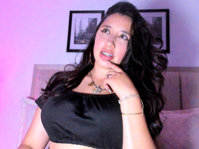 JessyBarnnett with long black hair, wearing a black crop top, holding her finger to her slightly open mouth, showing her teeth, and wearing a necklace and bracelet.