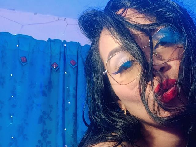 FetishLoverAna, con i capelli neri spettinati, rossetto rosso, ombretto blu e occhiali, che fa la smorfia mostrando le sue labbra lucide. Indossa orecchini dorati davanti a una tenda blu..