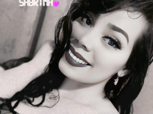 Das Gesicht von FetishLoverAna mit dunklem Make-up, weißen Zähnen und einem Lächeln, schwarzen Haaren, einem rosa Ohrring und einem grauen Hintergrund. Die Brüste sind nicht sichtbar, der Fokus liegt auf ihrem Gesicht..