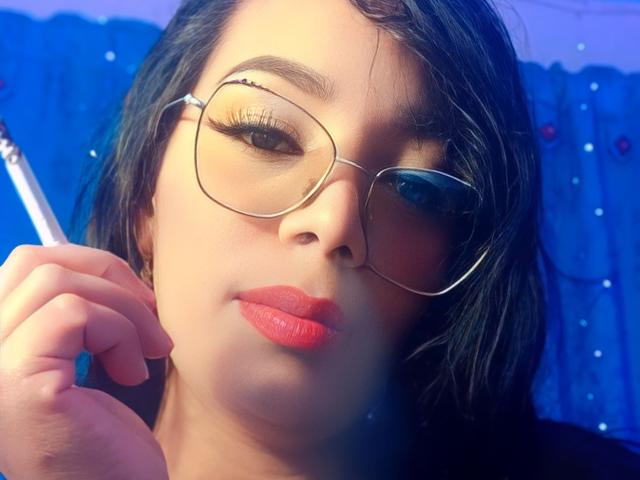 FetishLoverAna has dark hair, wears large glasses, and holds a pen near her face with red lipstick, looking at the camera with a slight smile.