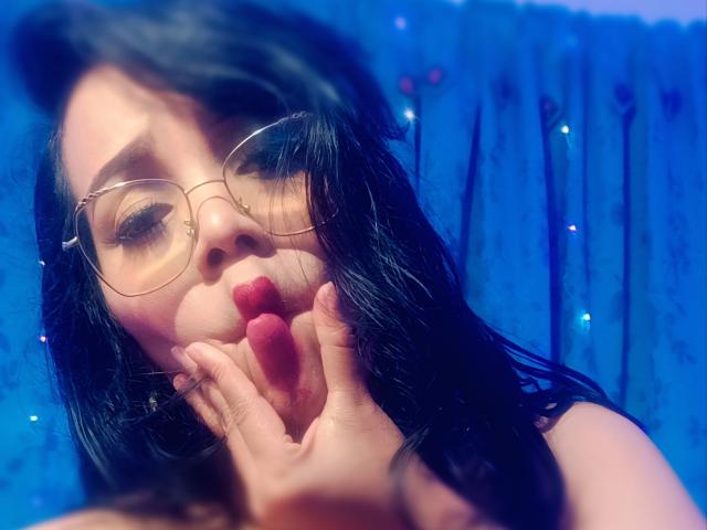 FetishLoverAna with black hair, wearing glasses, pouty red lips, and hand near face, blue background, slightly blurry, looking down, soft lighting, cute, sexy, focused on mouth.