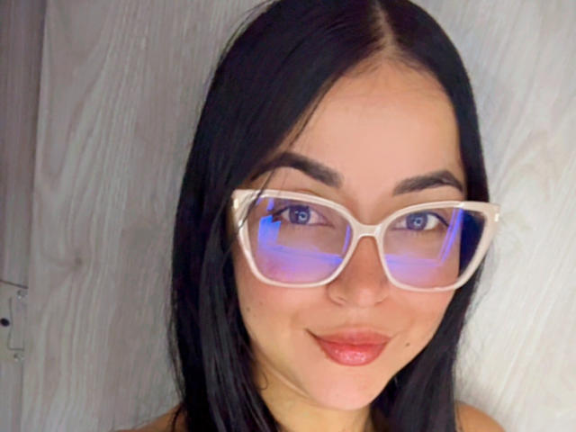 HikariChloe, avec ses cheveux noirs et lisses, portant des lunettes blanches avec un reflet bleu, et du rouge à lèvres, souriant à l'appareil photo..