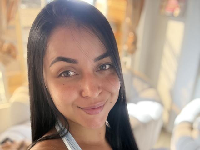 A HikariChloe tem cabelos pretos longos, pele clara, e está sorrindo, com seios pequenos, olhos castanhos e um nariz fofo. Ela está vestindo uma blusa branca de alças..