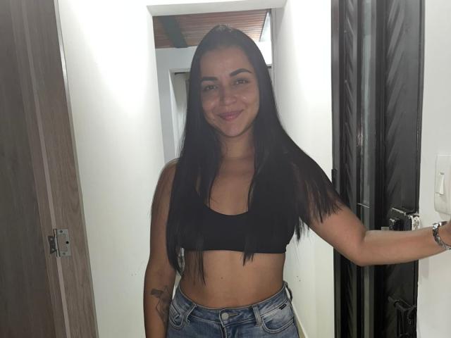 HikariChloe, com cabelos pretos longos, sorrindo, vestindo um top esportivo preto e jeans azuis, mostrando uma tatuagem no braço direito, em pé em uma porta..