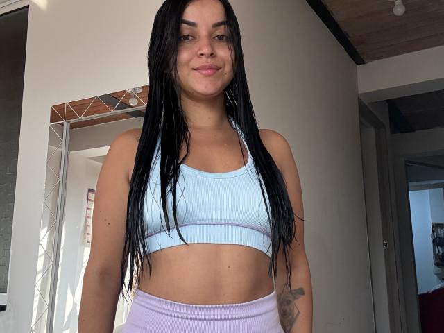 HikariChloe, com cabelos pretos longos, um top esportivo azul claro, mostrando seios pequenos, e calças roxas, em pé, sorrindo, com uma tatuagem no antebraço direito, pele clara, em ambiente interno, durante o dia..
