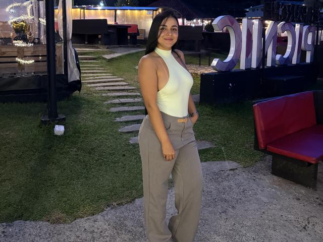 HikariChloe tiene un trasero curvilíneo, pechos de tamaño medio y cabello largo y oscuro. Lleva un top de tirantes amarillo y pantalones grises, y está de pie al aire libre por la noche, junto a un banco rojo..