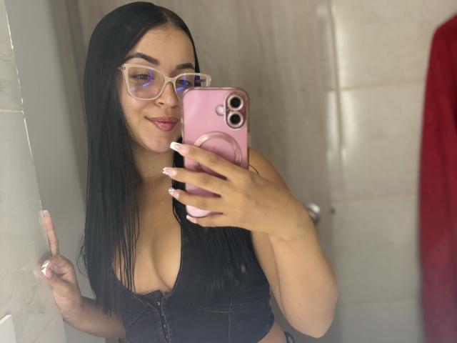 HikariChloe, com cabelos pretos longos, óculos e um telefone rosa, exibindo o decote em uma blusa preta com zíper, fazendo um sinal de paz. Seios, óculos, telefone rosa..