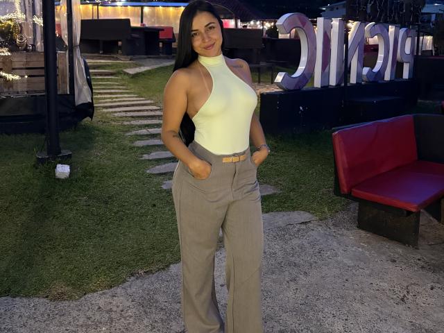 HikariChloe con cabello largo y negro, un top amarillo de tirantes que muestra sus pechos, y pantalones grises, con las manos en los bolsillos, sonriendo, de pie sobre la hierba con un banco rojo a la derecha..