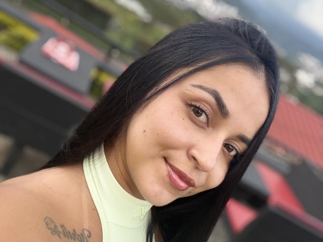 HikariChloe, con cabello negro largo, un top halter verde claro y un tatuaje pequeño en el hombro izquierdo, sonriendo a la cámara, ojos marrones, piel marrón clara, maquillaje sutil, lápiz labial rosa..