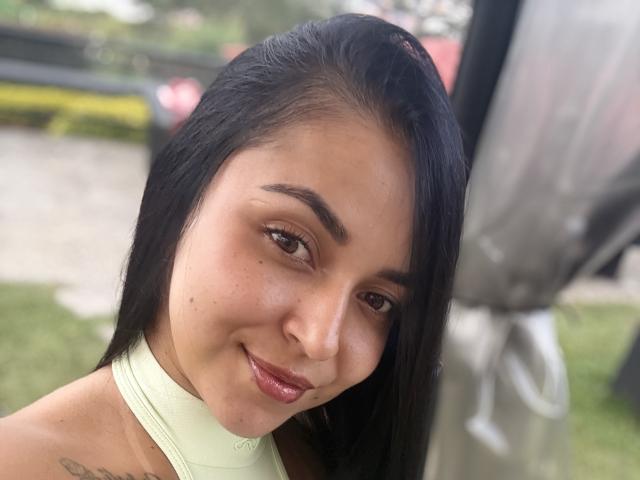 HikariChloe, com pele marrom média, cabelo preto e liso, e um pequeno sorriso, mostrando seus lábios vermelhos brilhantes, vestindo uma blusa verde clara com alças e uma pequena tatuagem no ombro..