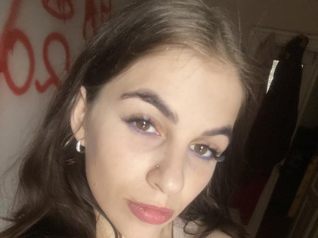 TrissyMima tem pele clara, olhos castanhos, delineador roxo, sobrancelhas escuras, um piercing no nariz e batom rosa. Ela está usando pequenos brincos de argola e tem cabelos castanhos longos..