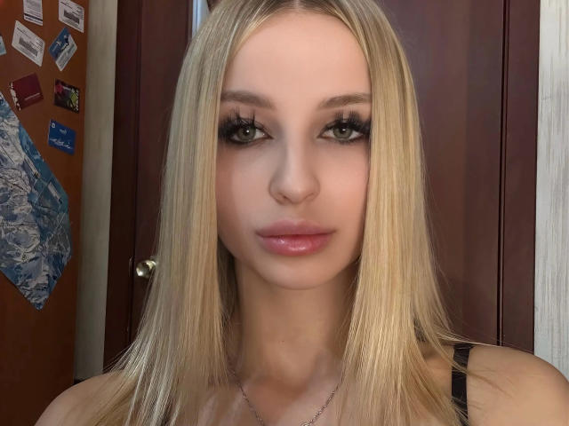 KeshaAites has long blonde hair, green eyes with thick black eyelashes, glossy pink lips, and is wearing a black top with a silver necklace.