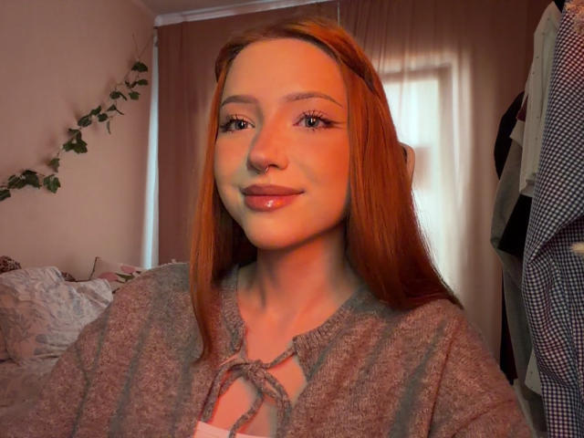 HeidyMerlette tiene el pelo rojo, un piercing en la nariz y lleva un suéter gris con un nudo atado. Está sonriendo con labios brillantes, luciendo linda y atractiva..