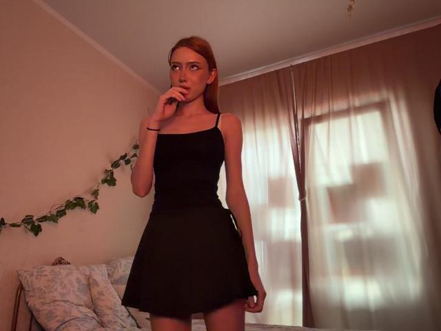 HeidyMerlette, con i capelli rossi, indossa un vestito nero a spalline sottili, è in piedi in una stanza, si tocca le labbra con un dito, guarda di lato, mostrando il suo sedere esile e minuto..