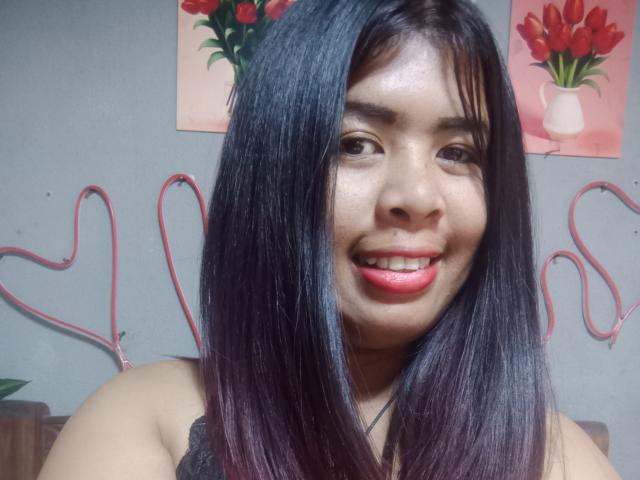 MariahSquirting69 ha i capelli neri e lisci, un naso piccolo e un sorriso con rossetto rosso. Indossa una maglietta nera, e sullo sfondo si vede un filo rosso a forma di cuore..