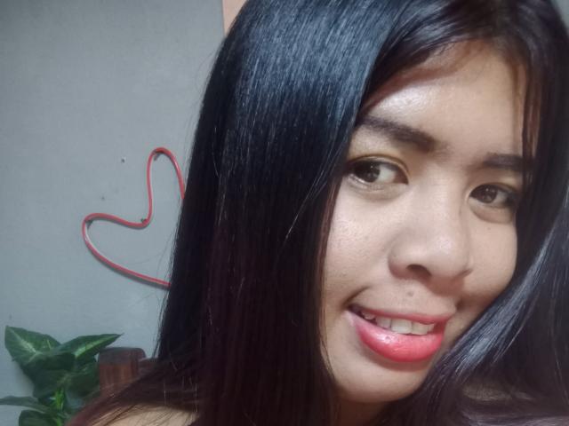 MariahSquirting69 con capelli neri, occhi marroni, rossetto rosa, che sorride, mostrando il suo viso, e un piccolo cuore disegnato in rosso su una parete grigia dietro di lei..