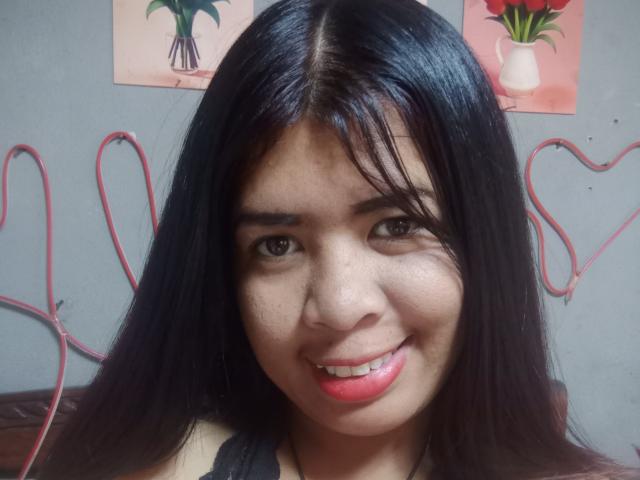 MariahSquirting69 ha lunghi capelli neri, occhi marroni e un leggero sorriso con rossetto rosso, mostrando un viso carino con qualche lentiggine, e indossa una maglietta nera..