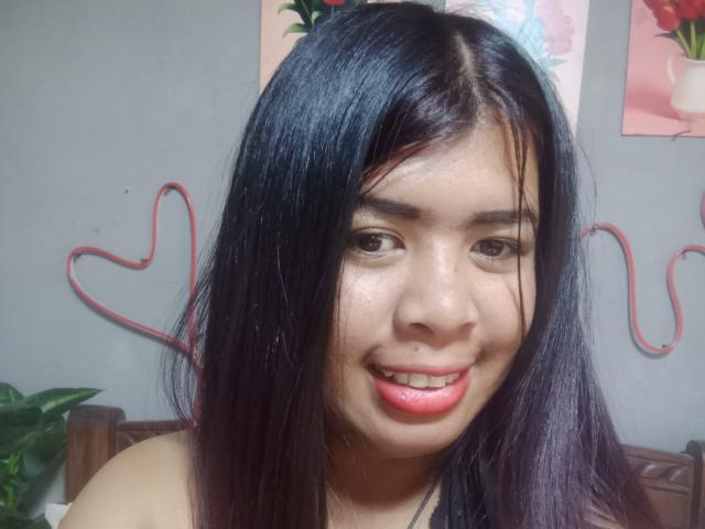 MariahSquirting69, con i capelli neri lucidi, gli occhi marroni e il rossetto rosso, sorride alla fotocamera, mostrando il piercing al naso e un viso leggermente lucido. Decorazione a forma di cuore rosso in filo metallico..