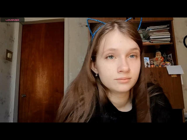 JessicaChrystal com orelhas de gato azuis, pele clara, cabelo castanho, olhos azuis e uma camisa preta, mostrando uma expressão neutra, em um quarto com uma porta de madeira e uma prateleira..