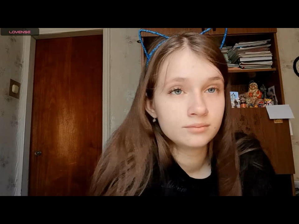 JessicaChrystal com orelhas de gato azuis, pele clara, cabelo castanho, olhos azuis e uma camisa preta, mostrando uma expressão neutra, em um quarto com uma porta de madeira e uma prateleira..