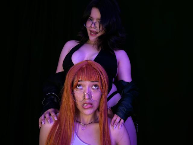 AlexaNDRoss, avec des cheveux foncés, un haut noir et une veste, assise derrière AlexaNDRoss, avec des cheveux orange, des lunettes et un haut blanc, les mains sur les fesses d'AlexaNDRoss aux cheveux orange, éclairage violet et bleu..