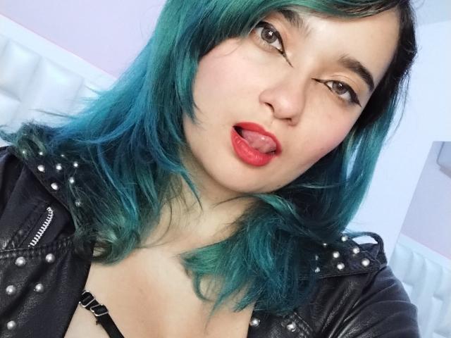 Naommy Carter, con i capelli verde acqua, una giacca di pelle nera e rossetto rosso, con la lingua fuori, che guarda la fotocamera, mostrando un reggiseno nero, carina e sexy, con un leggero ammiccamento..