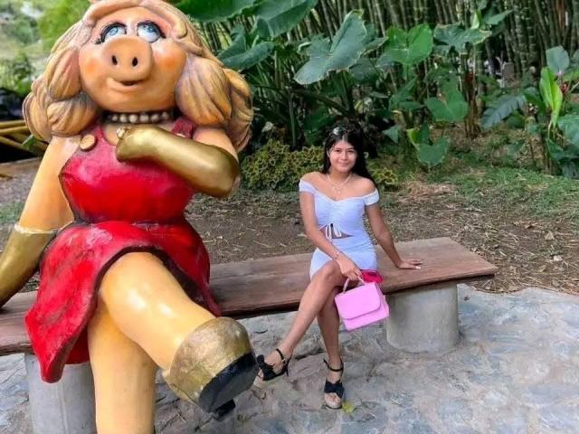 AshlyLee mit braunen Haaren, die ein weißes Kleid mit freiliegenden Schultern und schwarze High Heels trägt, sitzt auf einer Bank und hält eine pinke Handtasche. Neben ihr steht eine riesige, gelb-rote Schwein-Statue mit goldenen Stiefeln..