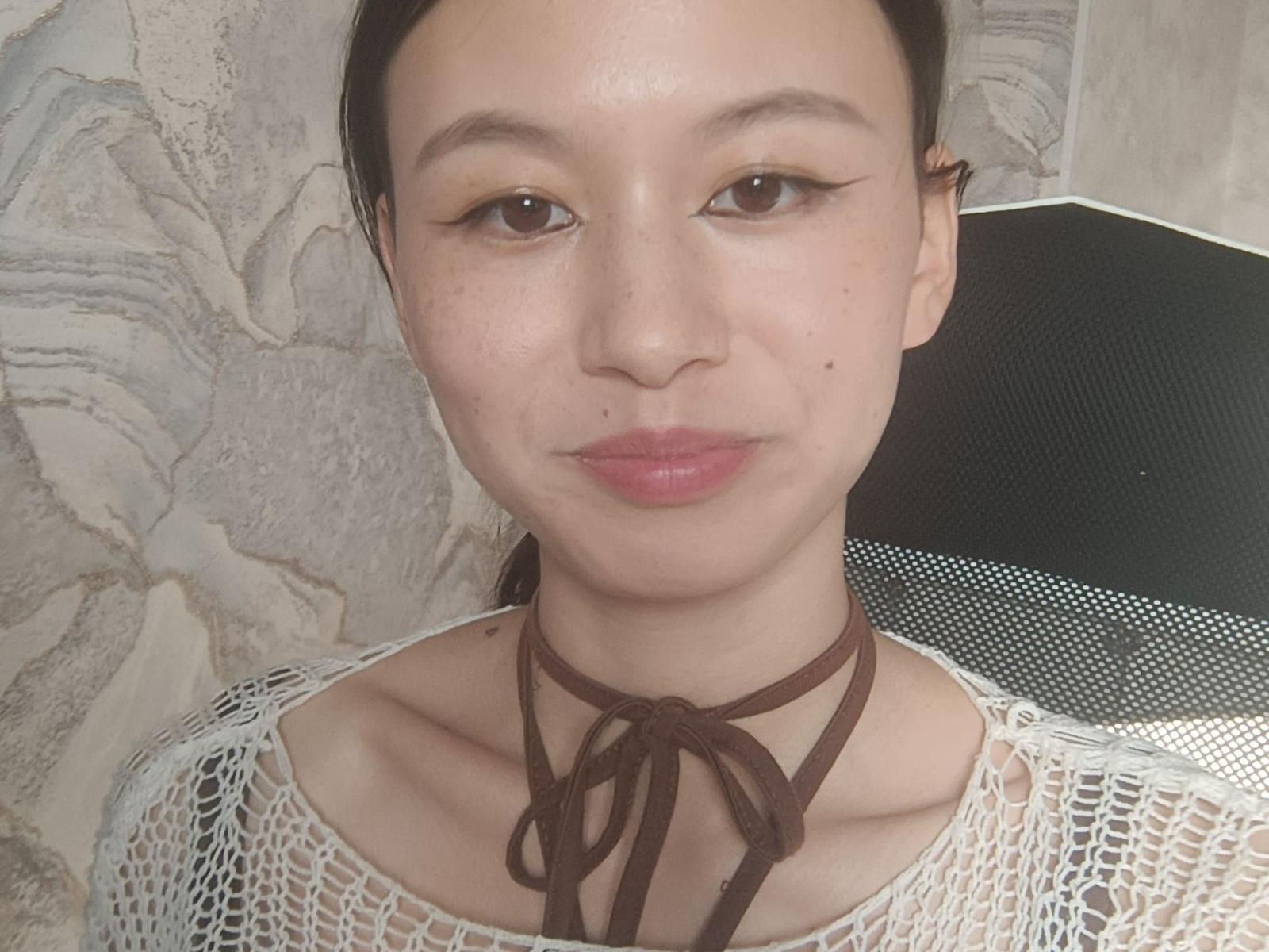 Emilia Brackemyre tem um rosto adorável, com um nariz pequeno e lábios rosados, usando um colar marrom e uma blusa branca de renda, olhos castanhos e uma pequena marca de beleza na bochecha direita..