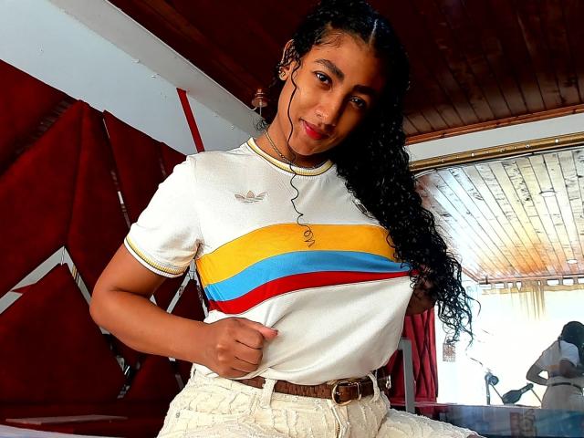 NatalyBlonder with curly black hair, white t-shirt with yellow, blue, and red stripes, and beige high-waisted pants, holding shirt, brown belt, wooden ceiling, red curtains.