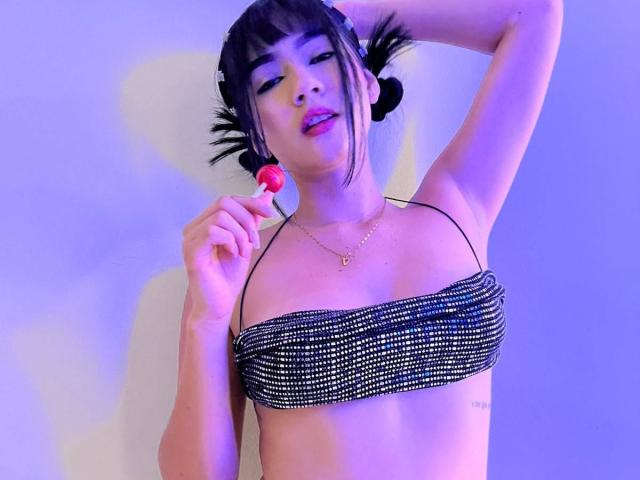 NahomiMillerr, con cabello negro en coletas, vistiendo un top negro brillante, sosteniendo una piruleta roja, levantando su brazo derecho y mirando a la cámara..