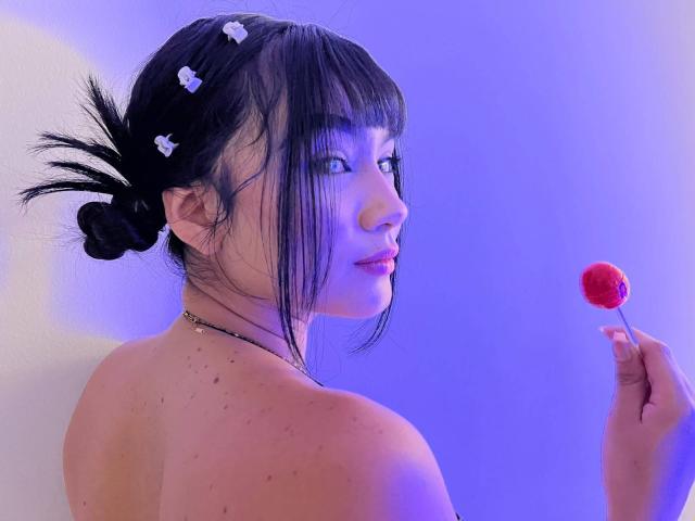 NahomiMillerr, avec des cheveux noirs attachés en chignons, des clips en forme de fleurs blanches, tenant un bonbon rouge, regardant en arrière, épaules nues, lumière bleu-violet, sourire doux, taches de rousseur, collier fin, expression douce..