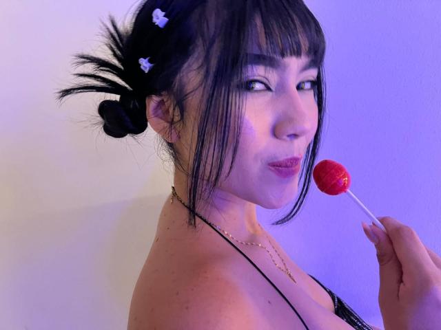 NahomiMillerr, avec des cheveux noirs attachés en chignon, des plumes noires, tenant un bonbon rouge près de ses lèvres charnues, portant un haut noir à bretelles et un collier en or..