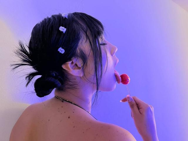 NahomiMillerr met zwart haar in een knot, met haarspelden, die een rode lolly zuigt, die ze vasthoudt met haar rechterhand, waarbij haar schouder en sproeten zichtbaar zijn, in een paars verlichte omgeving..