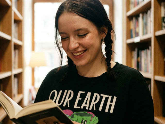 CandaceNotaro, sorridente con gli occhi chiusi, ha i capelli castani intrecciati, indossa una maglietta nera con la scritta "OUR EARTH" e sta leggendo un libro in una biblioteca, tenendo una tazza verde nella mano destra..