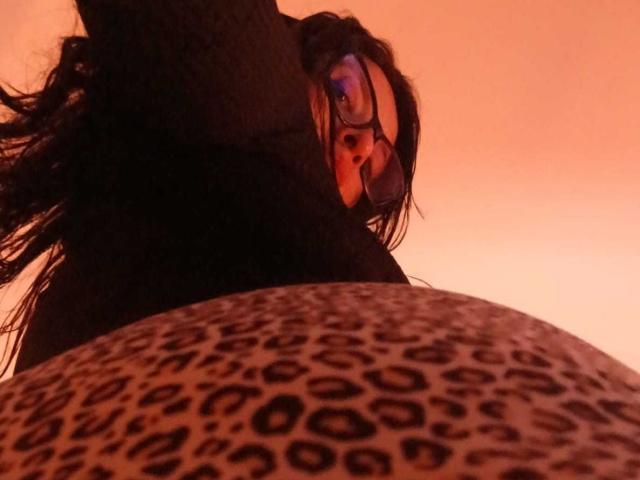 ZarayFeet with glasses, looking down, has long dark hair, wearing a black top, against an orange glow, with a leopard-printed object in the foreground.