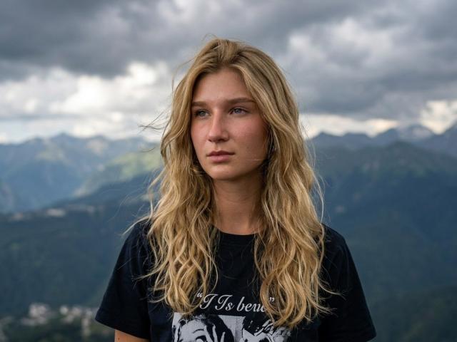 JinaKulakowski with long, wavy blonde hair, wearing a black t-shirt, standing outdoors, cloudy sky, mountain range in background, looking serious, slight pink in her cheeks.