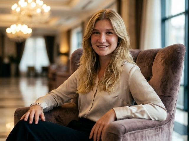 Jina Kulakowski, met lang blond haar, lachend, zittend in een paarse fluwelen fauteuil, gekleed in een beige blouse en een zwarte rok, in een goed verlichte kamer met kroonluchters..