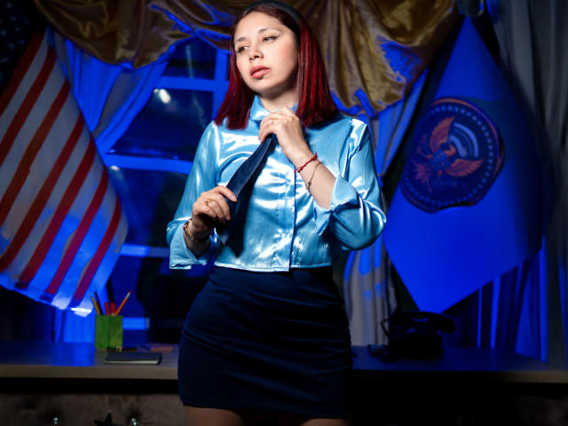 StephanieSun with red hair, blue shiny blouse, black skirt, holding black tie, looking confident, standing in front of American flag and Presidential seal. Her ass is cute.