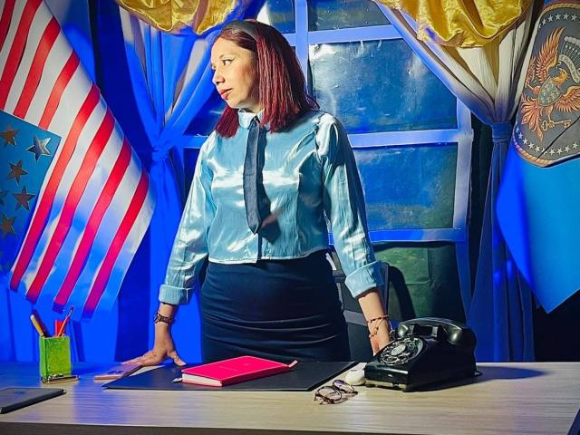StephanieSun with red hair, wearing a blue shirt and black skirt, standing in an office, hands on desk, black phone, red book, blue curtains, and American flag.