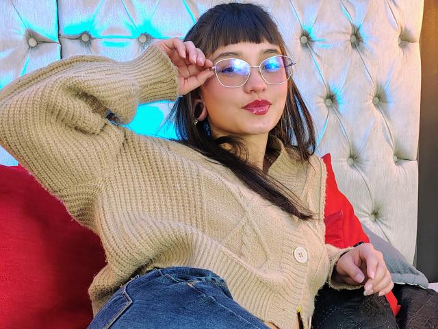 Alice Morely, con gafas, vestida con un suéter de punto beige y jeans azules, tiene un brazo levantado, mostrando sus pequeños senos, y está sentada sobre un cojín rojo..