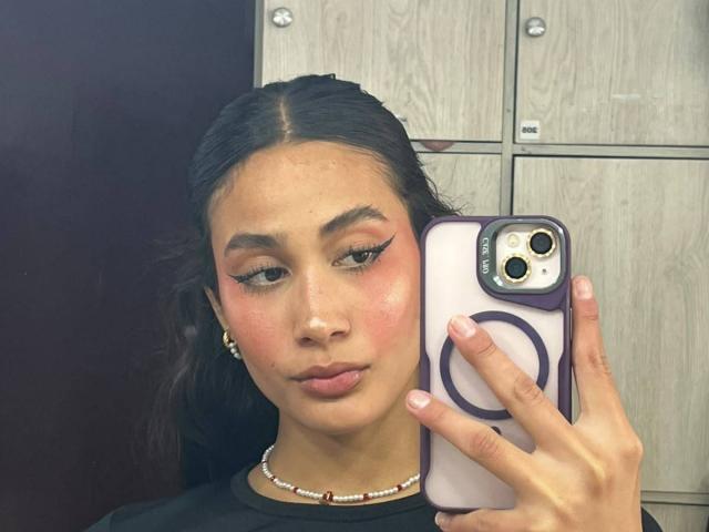 JennTylor with dark hair in a braid, pink blush, and winged eyeliner, holding a purple phone, pouting her lips, wearing a black shirt and bead necklace.