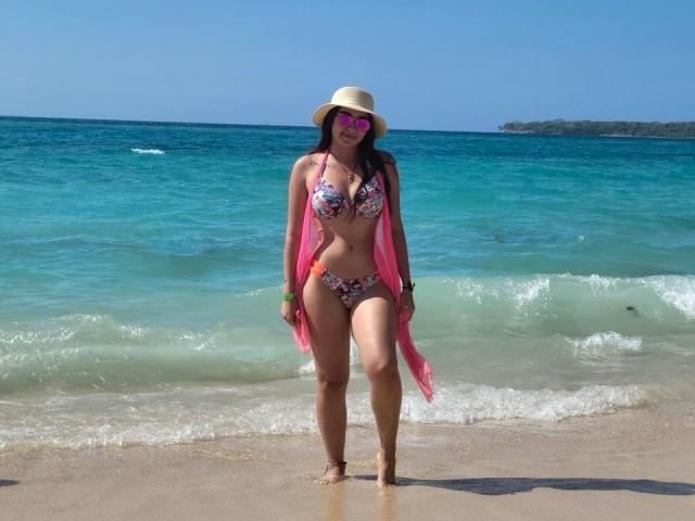 SalomeJoe in a floral bikini, pink scarf, sun hat, and pink sunglasses, standing on a beach with turquoise water, wearing a bracelet, smiling, with her ass and tits visible.