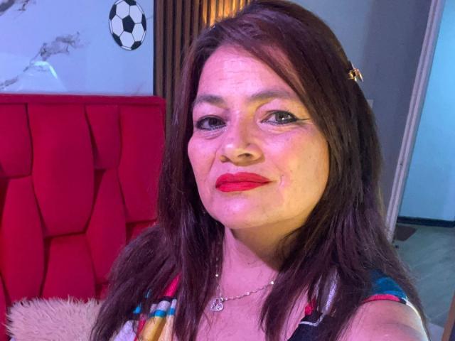 HannaMilff hat braunes, mittellanges Haar, roten Lippenstift und ein leichtes Lächeln. Sie trägt ein farbenfrohes Oberteil mit einer Halskette, und ihre Augen sind leicht hängend. Ihr Hintern ist nicht sichtbar..