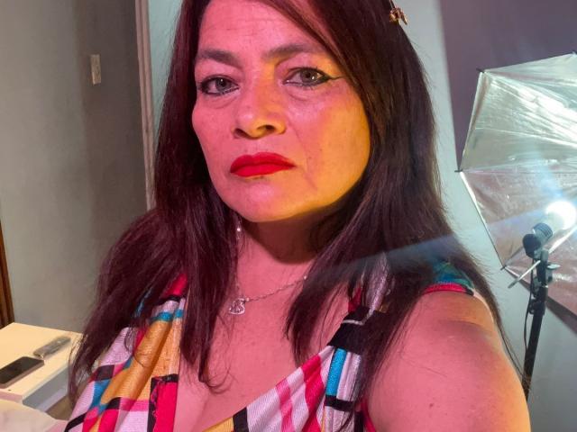 A HannaMilff tem batom vermelho, cabelo castanho e está vestindo um vestido colorido com estampas xadrez. Ela está olhando para a câmera com uma expressão neutra. Seus seios estão parcialmente visíveis..