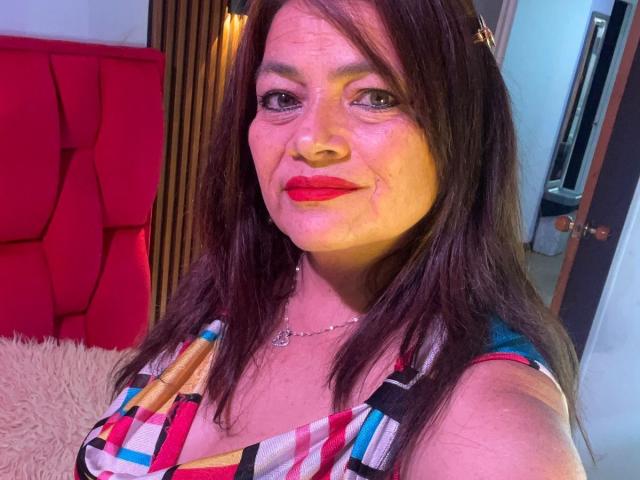 HannaMilff a les cheveux bruns, du rouge à lèvres, et une robe à carreaux. Elle sourit, ses yeux sont bleus, elle porte un collier et on voit des rides sur son visage. Elle tient un téléphone..