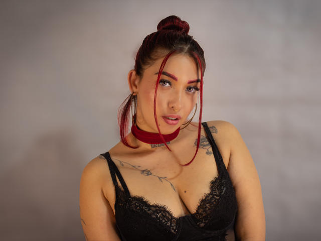 BriielaEva mit roten Haaren im Dutt, schwarzer Spitzen-BH, Tattoos auf der Brust, roter Choker, blickt in die Kamera, helle Haut, große Brüste, leichtes Lächeln, Hintergrund unscharf..