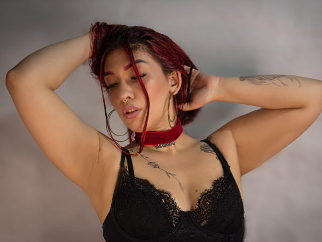 BriielaEva mit roten Haaren, einem schwarzen Spitzen-BH und einer roten Halskette, mit erhobenen Armen, geschlossenen Augen, einem Tattoo am linken Arm, einem Nasenring und Creolen, wirkt sie sexy und selbstbewusst..