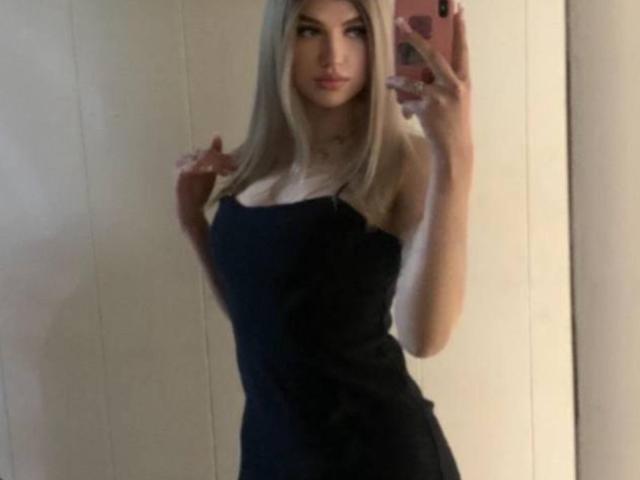 Tawanna Lehrman mit langen, blonden Haaren, trägt ein schwarzes, trägerloses Kleid, hält ein rosa Telefon, zeigt ihre Brüste, steht vor einem Spiegel und schaut in die Kamera..